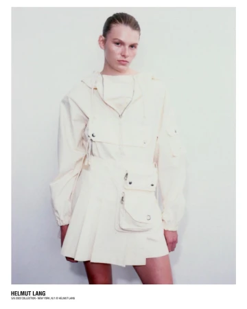 Model wearing a cream-colored utility jacket and pleated miniskirt against a plain light wall in a portrait by Dario Catellani.