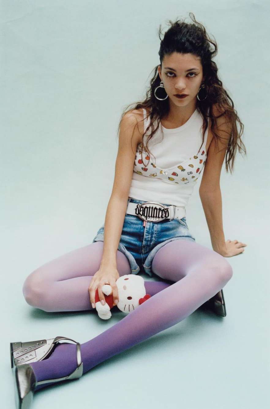 A woman sits in a studio with blue tights and jean shorts, holding a Hello Kitty plush, wearing a white printed top and silver shoes.