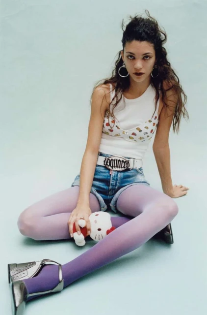 A woman sits in a studio with blue tights and jean shorts, holding a Hello Kitty plush, wearing a white printed top and silver shoes.