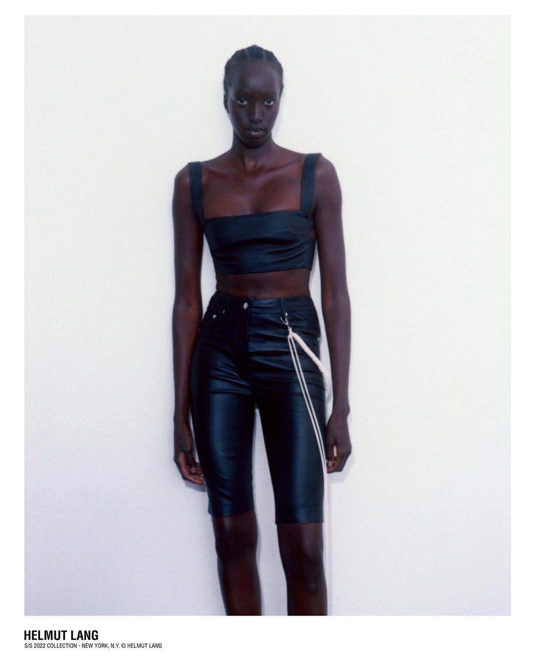 Fashionable woman in a black crop top and black shorts poses against a white wall in a studio setting, wearing Helmut Lang brand.