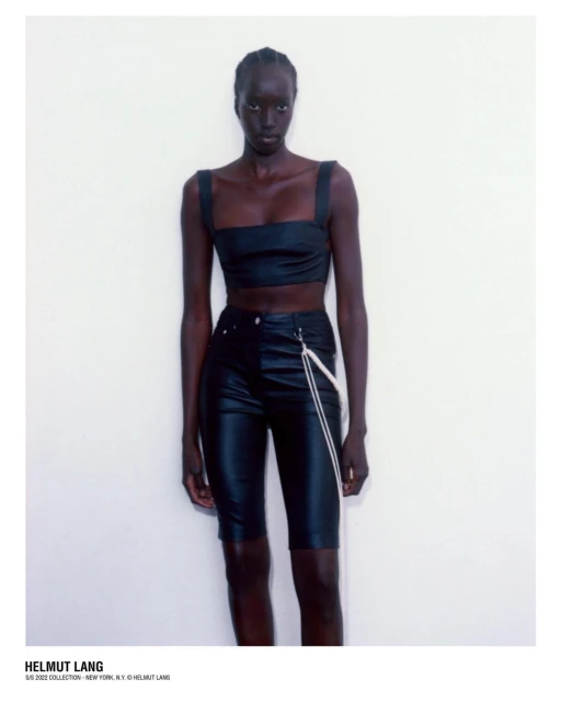 Fashionable woman in a black crop top and black shorts poses against a white wall in a studio setting, wearing Helmut Lang brand.