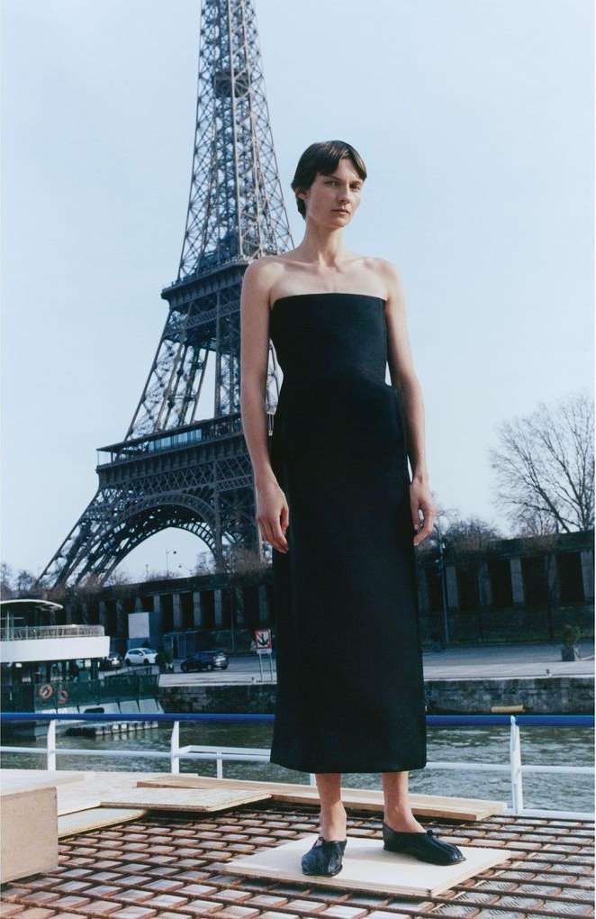 A lone woman in a black dress poses in front of the Eiffel Tower, Paris, under a bright sky, capturing a blend of fashion and iconic architecture.