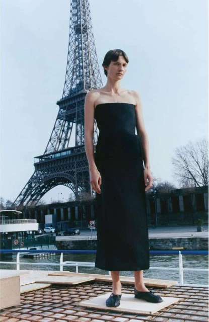 A lone woman in a black dress poses in front of the Eiffel Tower, Paris, under a bright sky, capturing a blend of fashion and iconic architecture.