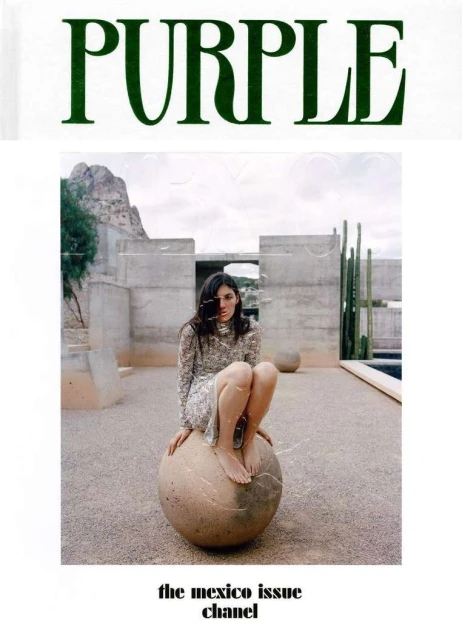 Fashion editorial shot featuring a woman with long dark hair in a floral dress, sitting on a round stone sculpture in an outdoor setting.