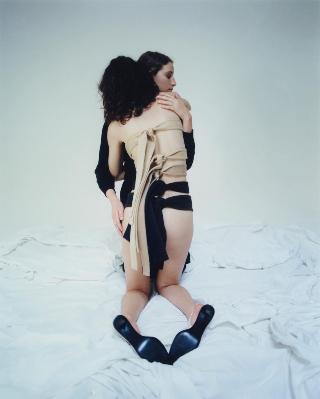 Two women embrace on a bed in a photo studio. One wears black heels and a wrapped garment, creating a minimalist and intimate portrait.