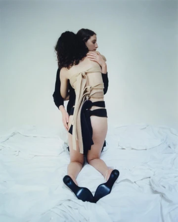 Two women embrace on a bed in a photo studio. One wears black heels and a wrapped garment, creating a minimalist and intimate portrait.