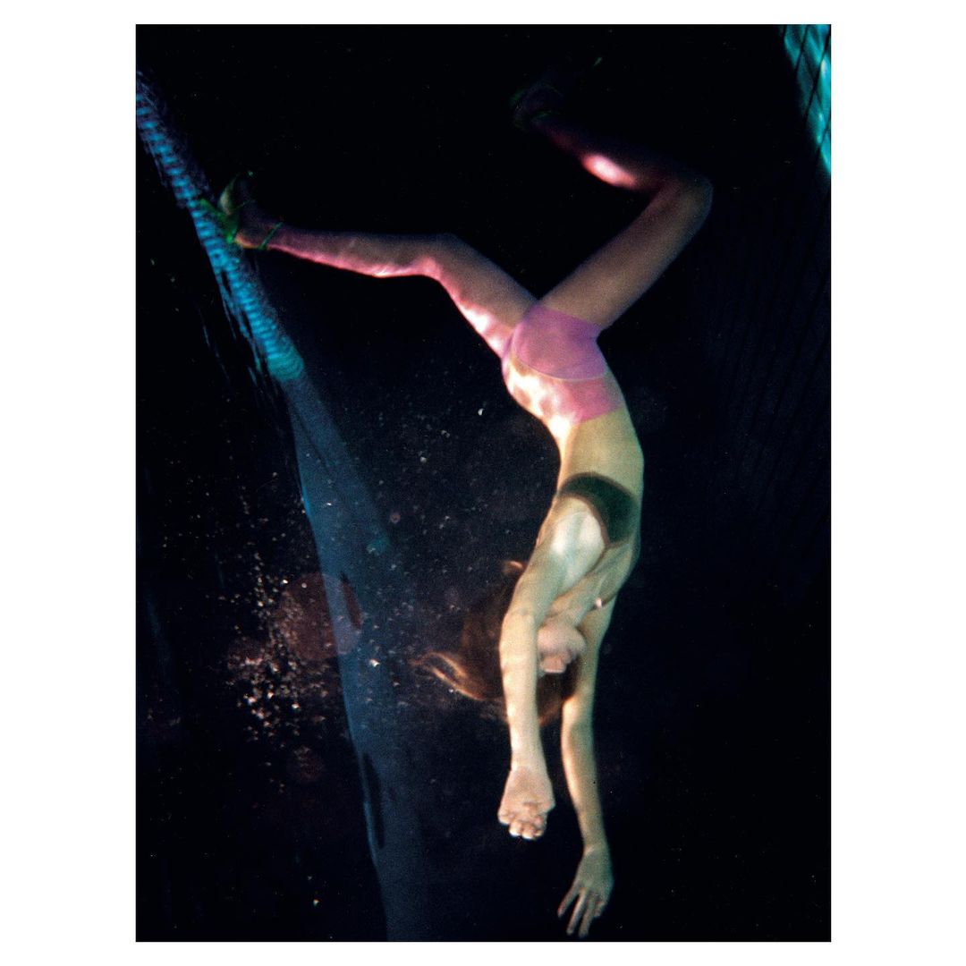 A person is upside down underwater, wearing pink shorts and a dark top. The individual appears to be performing a handstand in dark water.