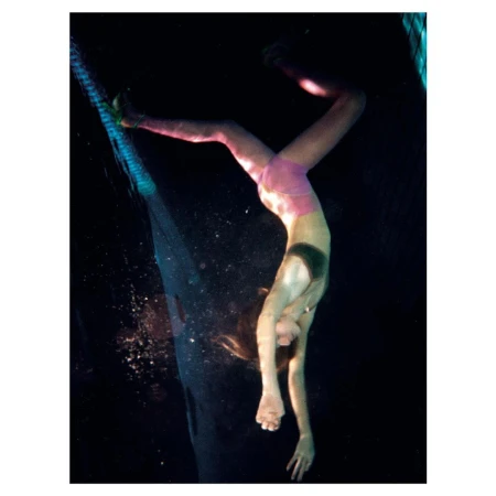 A person is upside down underwater, wearing pink shorts and a dark top. The individual appears to be performing a handstand in dark water.