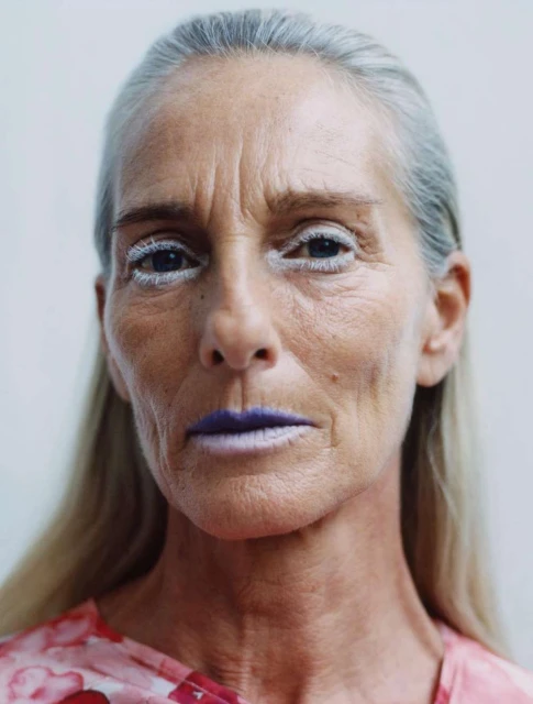Close-up portrait of an older adult female with fair skin, white hair, exaggerated white eye makeup and light purple lipstick on a white backdrop.