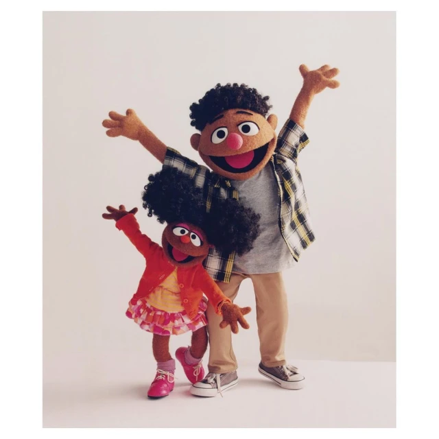 Two Sesame Street puppets, a girl and a boy, stand against a light backdrop with their arms raised in a cheerful pose, creating a playful image.