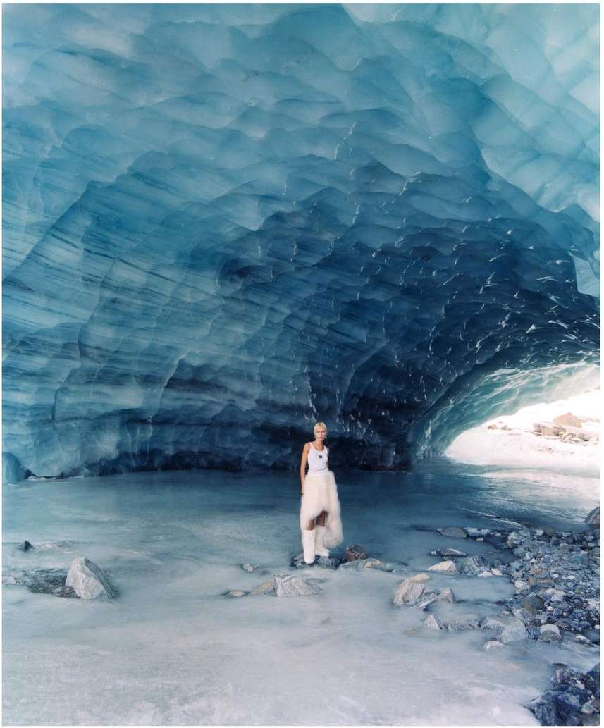Adult woman in a white dress standing in an ice cave with blue ice formations, water, and rocks, captured in a wide shot.