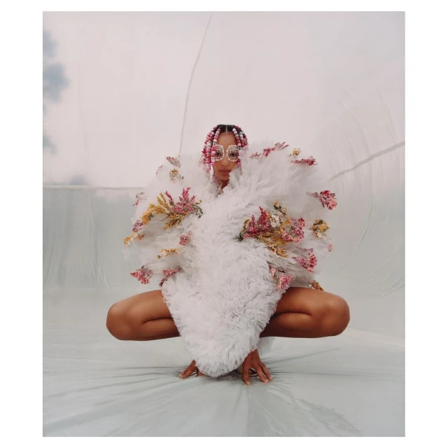 Woman crouching wearing a white ruffled floral gown and geometric sunglasses in a light studio environment. Fashion editorial photography.
