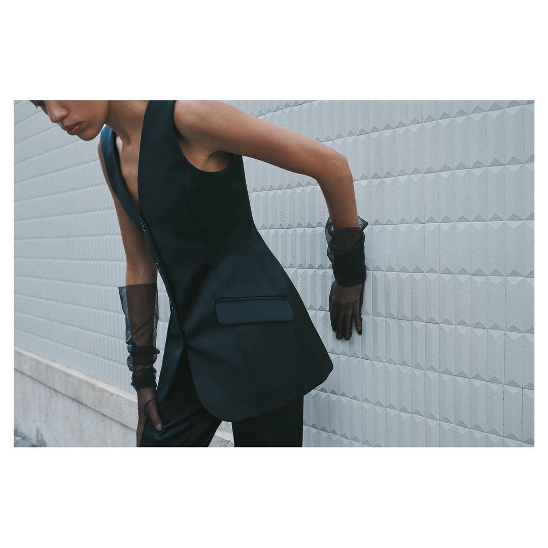 An adult female poses in a black vest and sheer black gloves against a textured grey wall, showcasing a minimalist and fashionable aesthetic.