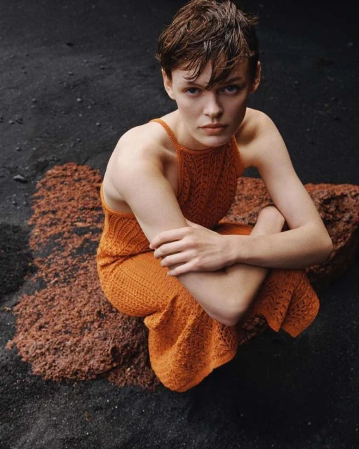 A young, fair-skinned adult woman with short brunette hair wears an orange knitted dress, kneeling on dark rocks in an outdoor setting.