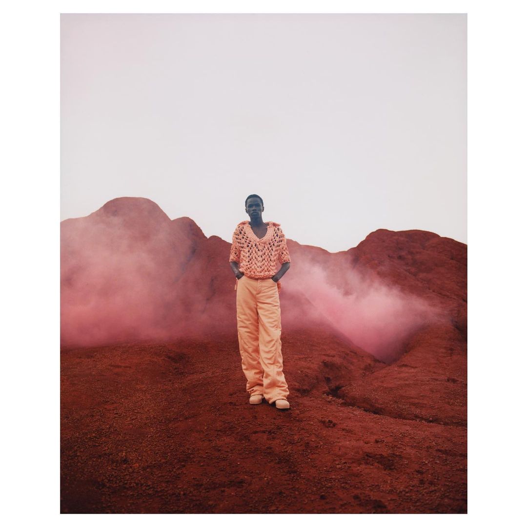 Woman in crocheted top and pants stands in a reddish-brown landscape with hazy pink smoke, under a pale sky, fashion-forward.