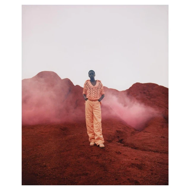 Woman in crocheted top and pants stands in a reddish-brown landscape with hazy pink smoke, under a pale sky, fashion-forward.