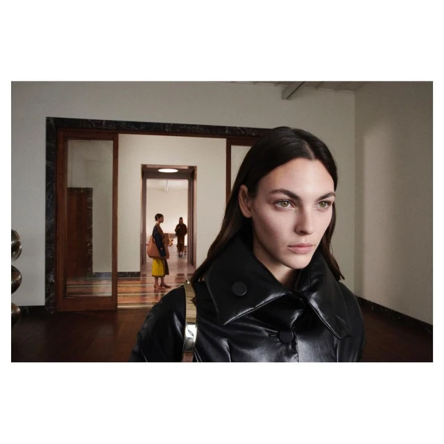 Elegant woman in a black jacket, posing indoors near an open doorway, with other people in the background; fashion editorial.