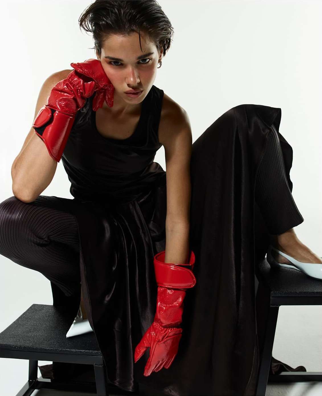 A fashion image features a young adult with short dark hair posing in a black tank top, dark pants, red gloves, and white heels against white.