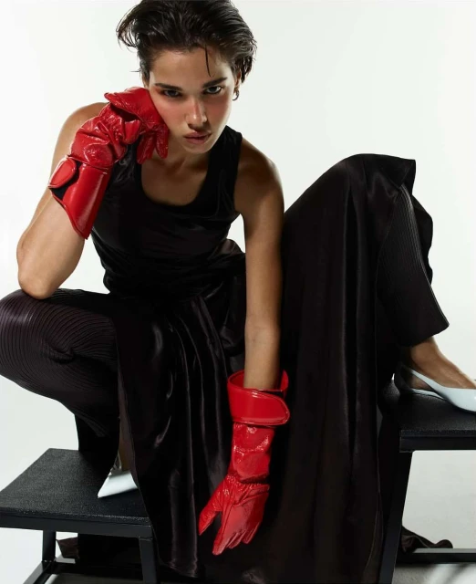 A fashion image features a young adult with short dark hair posing in a black tank top, dark pants, red gloves, and white heels against white.