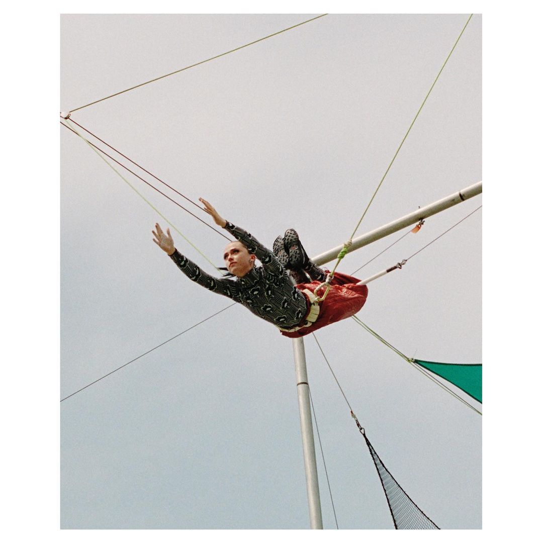 A person wearing a patterned bodysuit performs on a trapeze, suspended against a light sky, showcasing skill and athleticism in an outdoor setting.