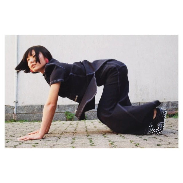Asian woman in dark pantsuit with contrasting stitching crawls on all fours on concrete in outdoor fashion shot against neutral background.