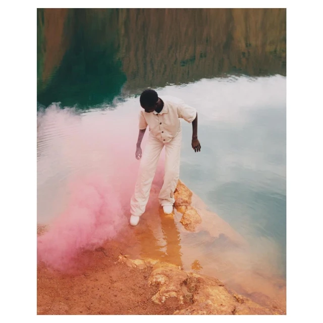 A person in a beige outfit stands on a rock near a body of water with a pink smoke bomb, creating a surreal and visually striking image.