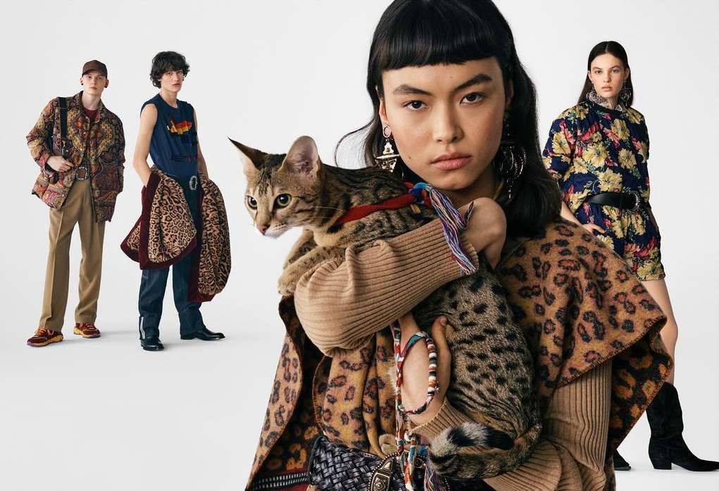 Fashion editorial image featuring a woman holding a cat, showcasing patterned clothing, with three additional adults in the background against a white backdrop.