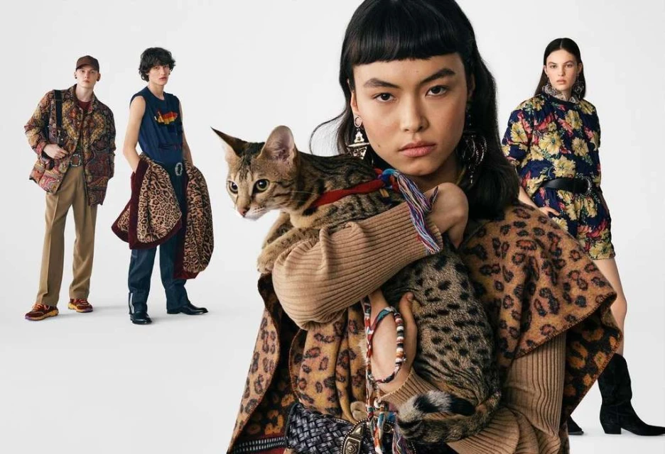 Fashion editorial image featuring a woman holding a cat, showcasing patterned clothing, with three additional adults in the background against a white backdrop.