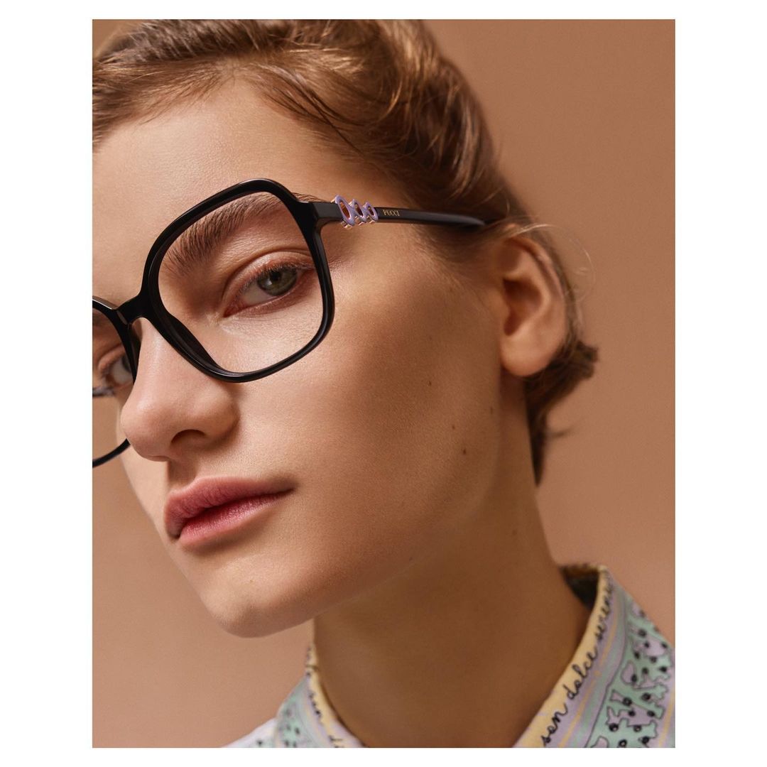 Close-up portrait of a woman wearing thick black frame eyeglasses and a patterned collar shirt against a solid brown background.