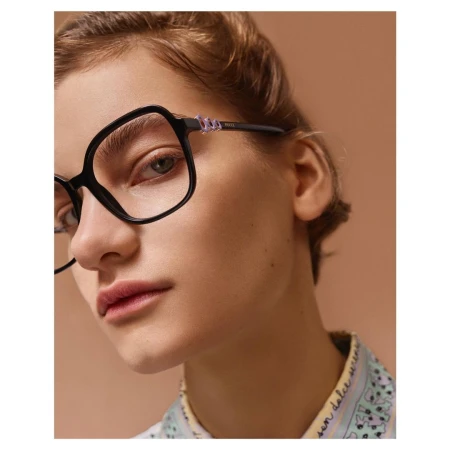 Close-up portrait of a woman wearing thick black frame eyeglasses and a patterned collar shirt against a solid brown background.