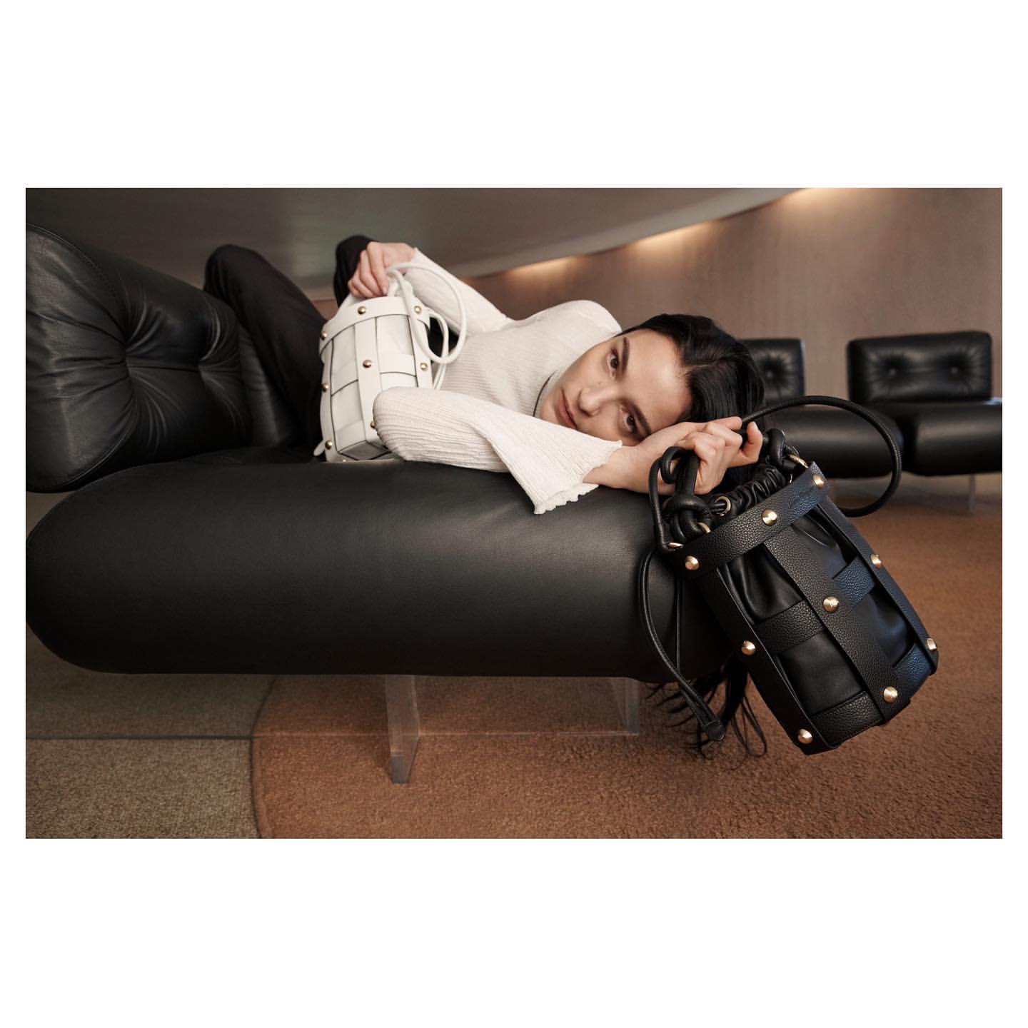 Woman poses with a black leather woven bucket bag and a white leather woven bucket bag, reclining on a black leather lounge.
