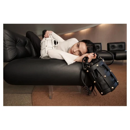 Woman poses with a black leather woven bucket bag and a white leather woven bucket bag, reclining on a black leather lounge.