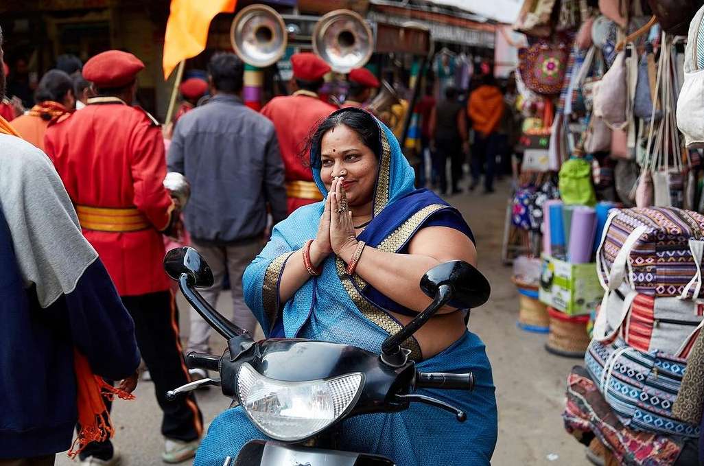 Adult woman in blue sari riding a scooter on a busy street, with a group of people in red uniforms and market stalls in the background.
