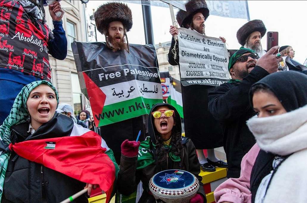 Large outdoor protest with diverse group of people holding signs and flags, chanting, in daylight. Fur hats and traditional headscarves.