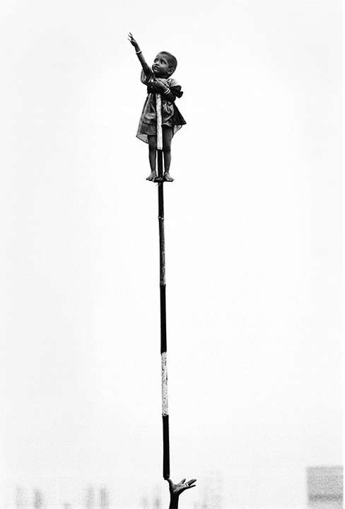 Black and white portrait of a child balancing on a pole and reaching up, with a hand reaching up from the bottom of the image.