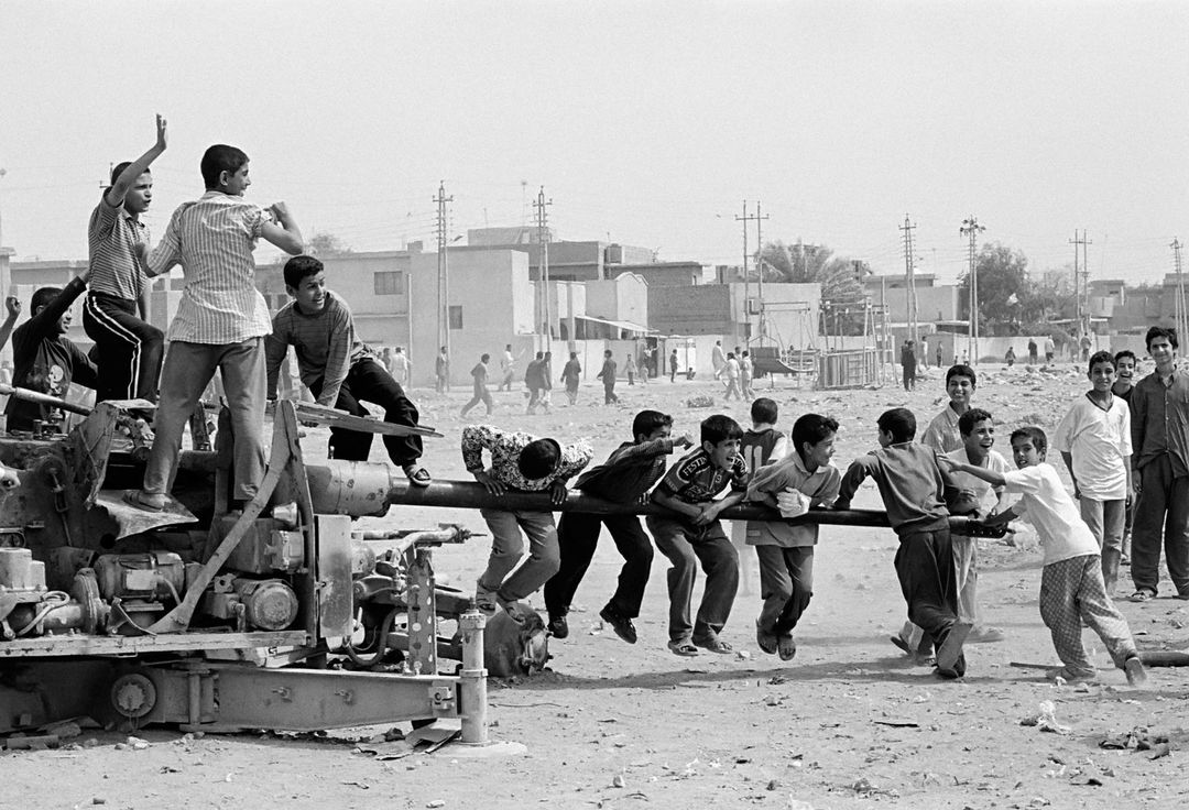 Black and white shot of a group of children in a war-torn area, climbing on a disused cannon, with buildings in the background.