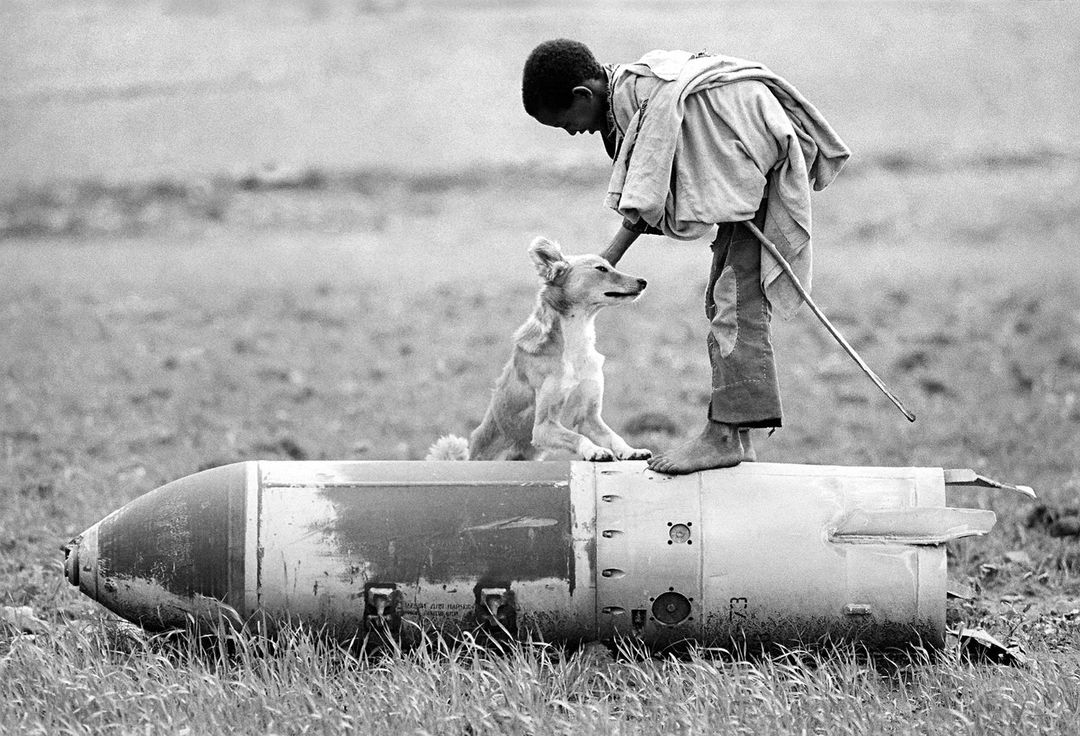 A child stands barefoot atop a warhead with a dog in a black and white outdoor scene, showing a poignant juxtaposition of innocence and conflict.