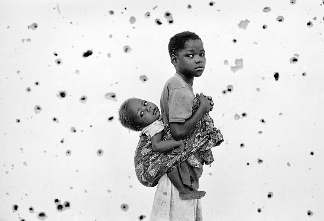 Monochrome portrait of an African child carrying a baby on their back, standing in front of a weathered wall with dark marks.