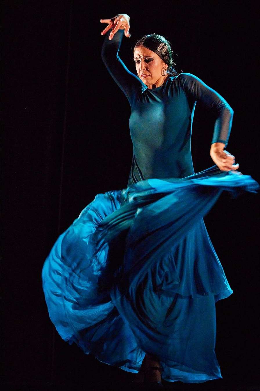 Female flamenco dancer in motion with traditional long blue dress against dark background, performing dance with raised arms in a studio setting.
