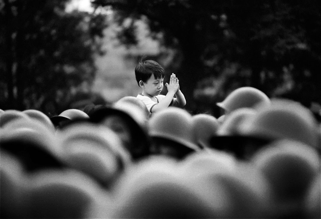 Black and white image shows a young boy with hands pressed together in prayer amidst a field of blurred army helmets.