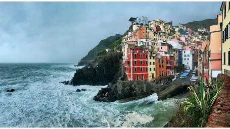 Coastal village with colorful stacked buildings perched on rocky cliffs above churning ocean waves under an overcast sky.