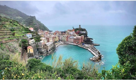 Coastal Italian village nestled around a turquoise blue bay with terraced green hills under an overcast sky.