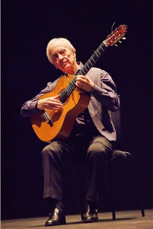 Elderly man playing acoustic guitar while seated on stage under dramatic lighting