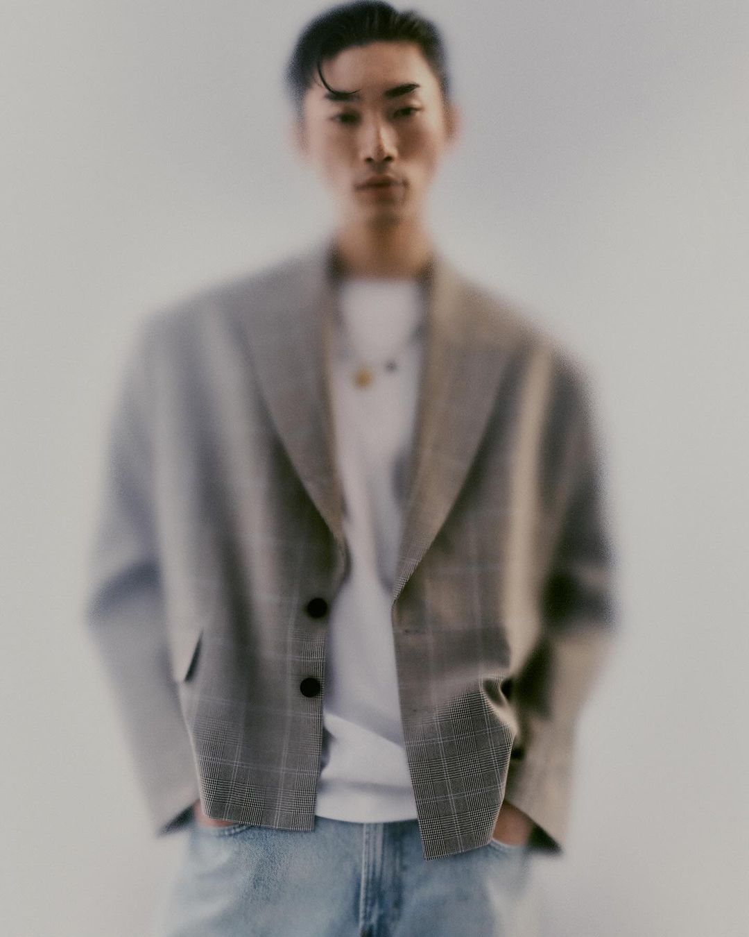 A blurry portrait of a young adult man wearing a grey plaid blazer over a white t-shirt and light blue jeans, posing indoors.