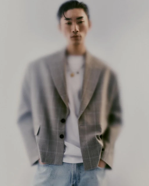 A blurry portrait of a young adult man wearing a grey plaid blazer over a white t-shirt and light blue jeans, posing indoors.