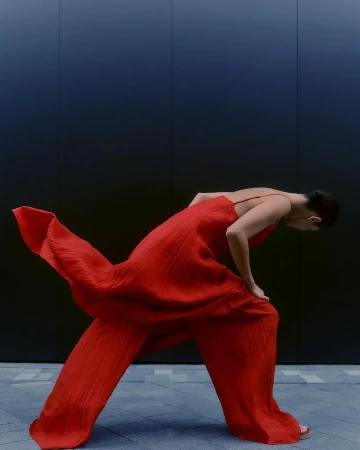 Model in a bright red textured jumpsuit leaning forward against a dark panelled wall outdoors in London.