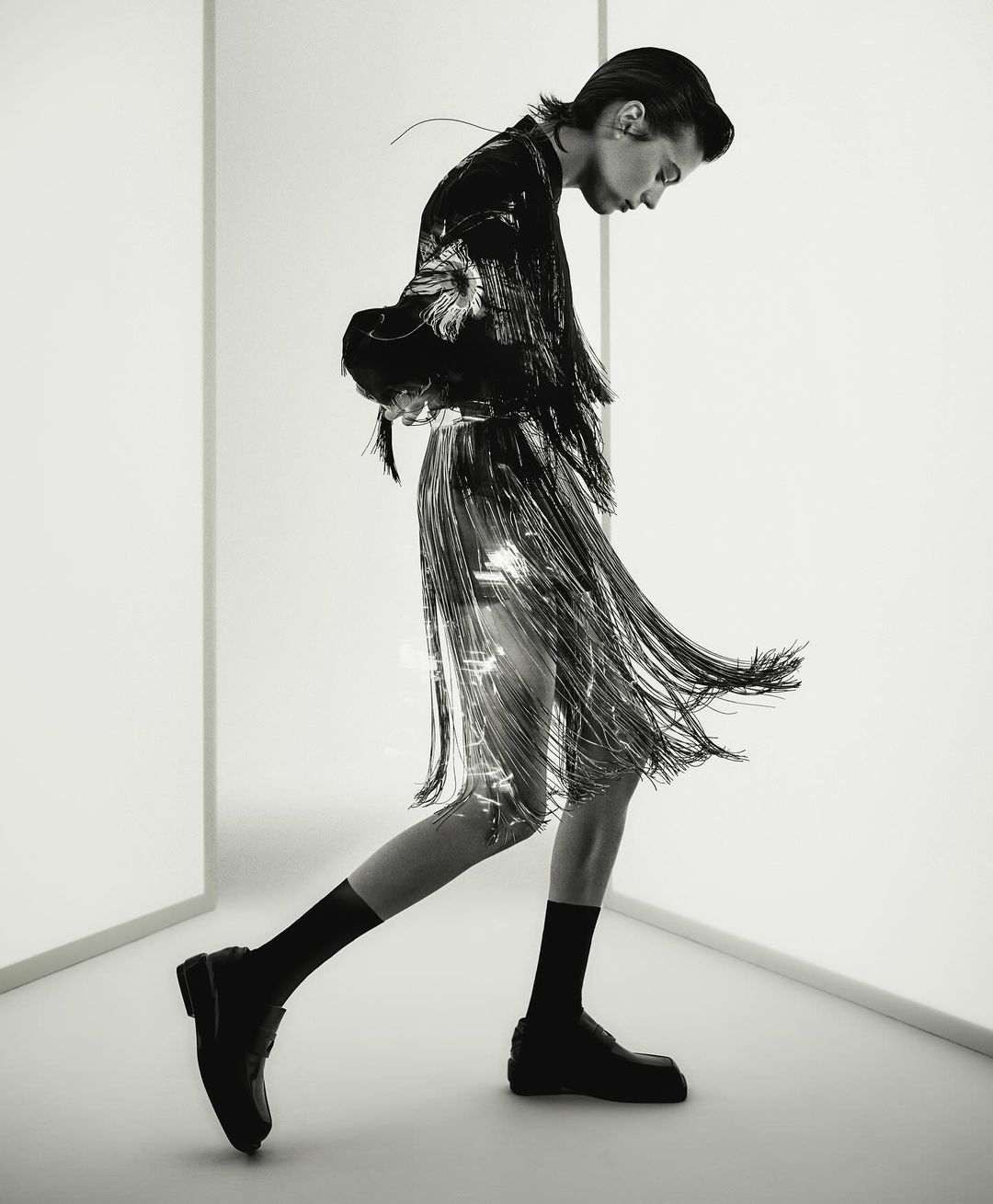 Fashionable adult in a shiny fringed skirt and black jacket walks in a white photo studio with black socks and chunky black loafers.