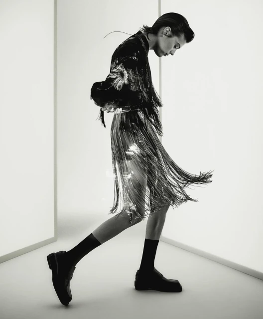 Fashionable adult in a shiny fringed skirt and black jacket walks in a white photo studio with black socks and chunky black loafers.