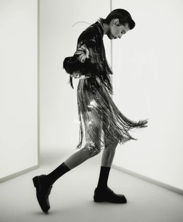 Fashionable adult in a shiny fringed skirt and black jacket walks in a white photo studio with black socks and chunky black loafers.