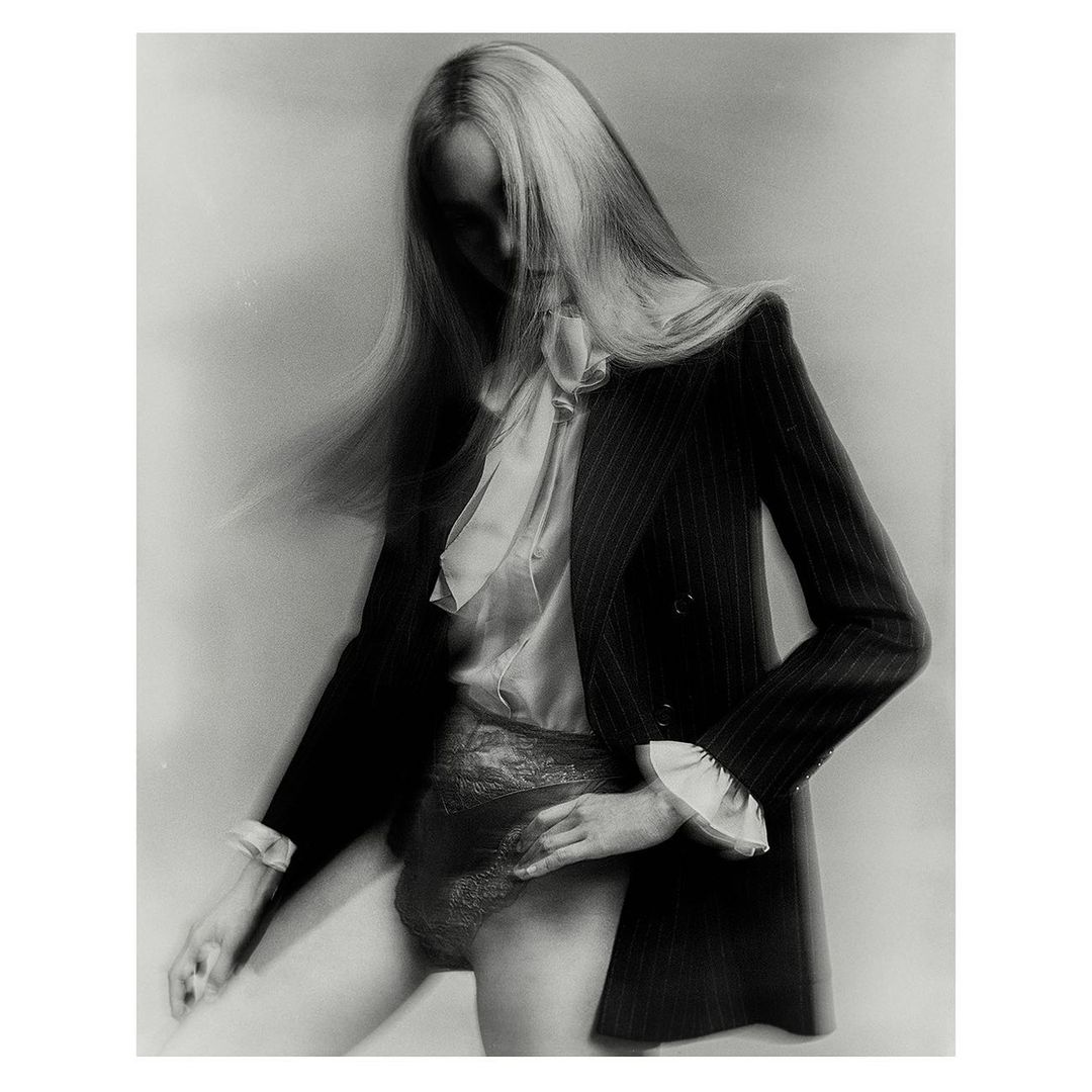 Monochrome portrait of a woman in a pinstripe blazer, blouse, and lace underwear against a simple light gray backdrop, capturing a minimalist fashion aesthetic.
